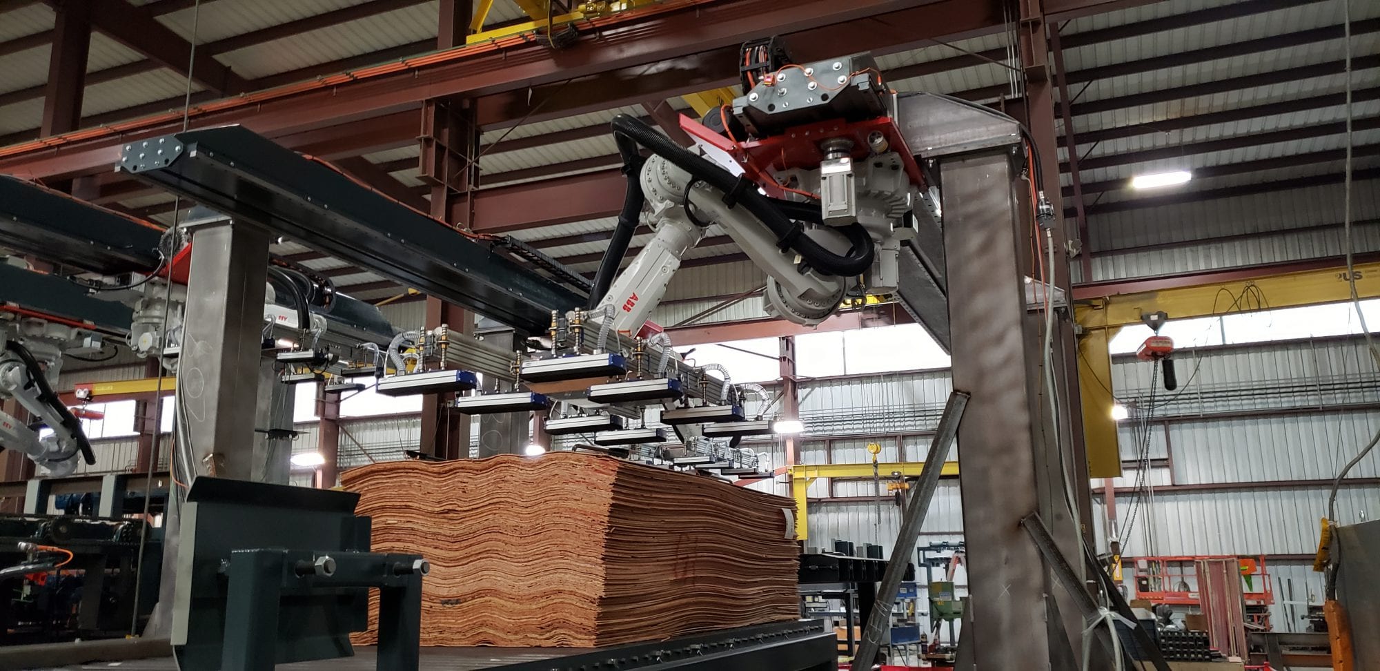 A robotic arm stacks thin wood sheets inside an industrial warehouse.