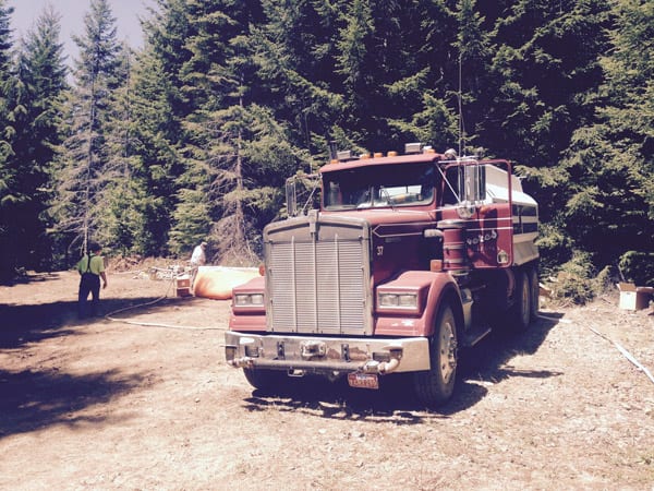 A red water tanker truck, marked with Freres Engineered Wood signage, is parked on a dirt clearing surrounded by tall pine trees.