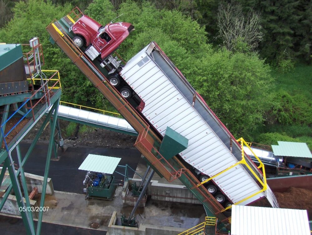 A semi-truck and trailer are tilted upward on a platform for unloading near a cogeneration industrial facility.