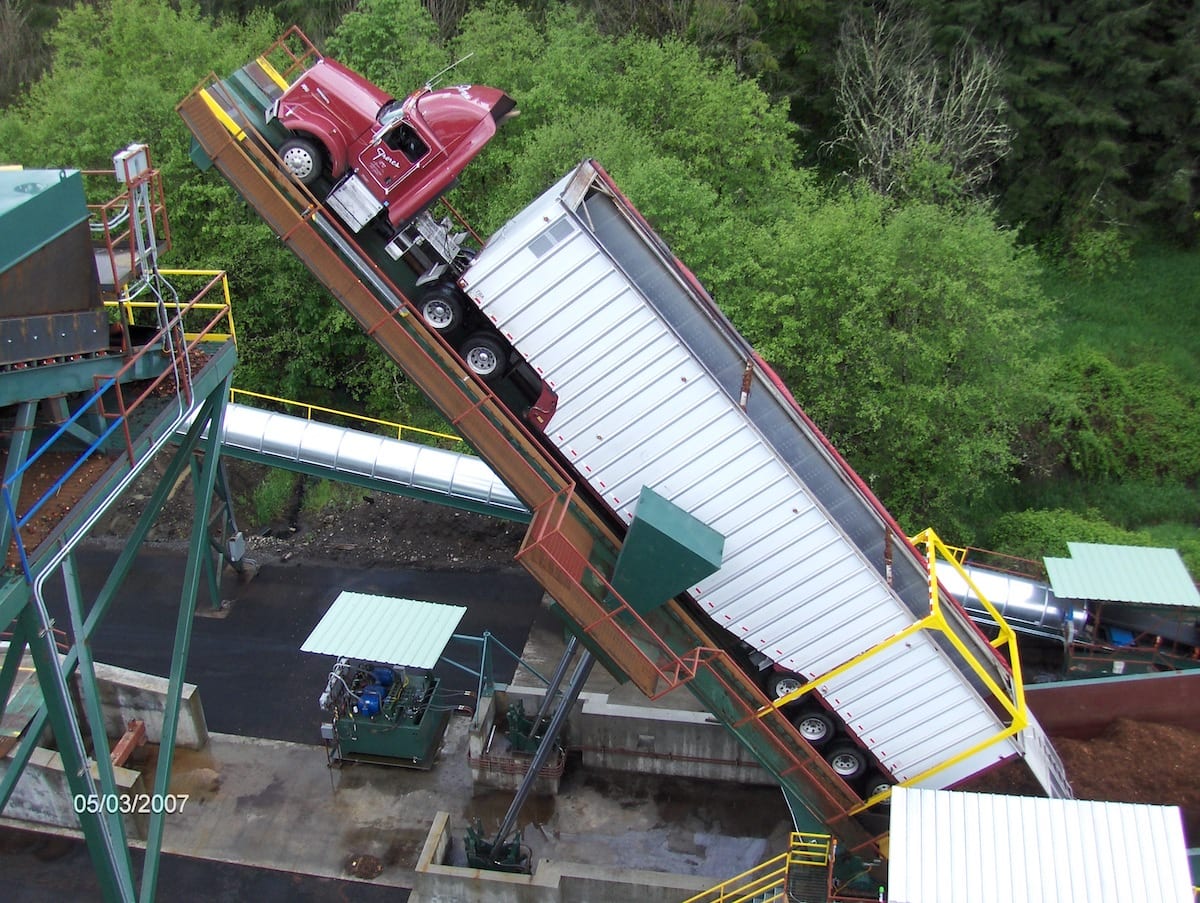 A semi-truck and trailer are tilted upward on a platform for unloading near a cogeneration industrial facility.