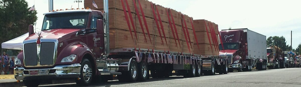 Several red semi-trucks hauling large stacks of plywood are lined up on a street during an event.
