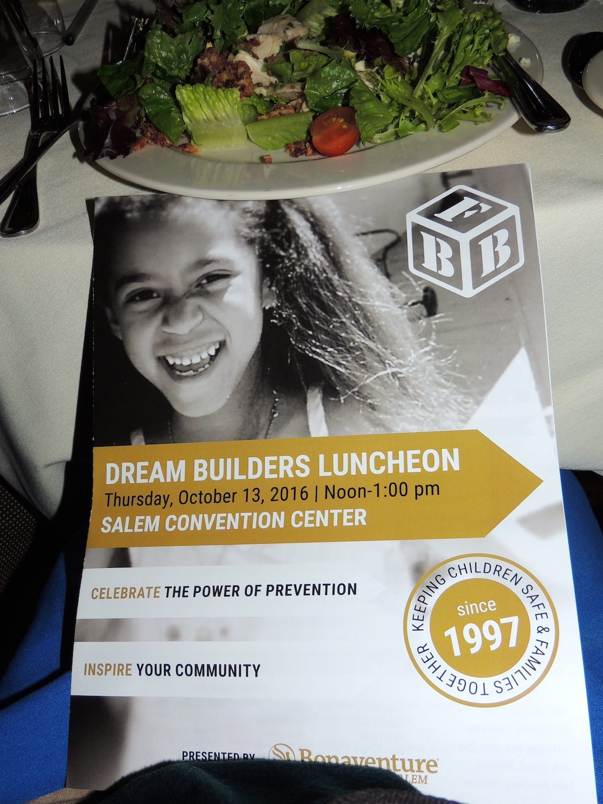 A flyer for Family Building Blocks' Dream Builders Luncheon 2016 rests on a table, with salad and utensils in the background.