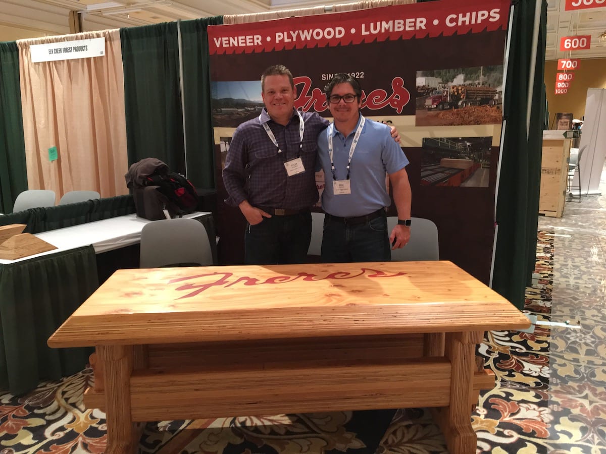 Two men stand behind a wooden table at a NAWLA Traders Market booth with a Freres banner.