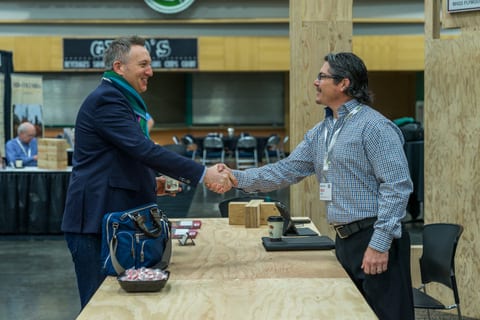 Two men in business attire shake hands across a wooden table at the Mass Timber Conference.