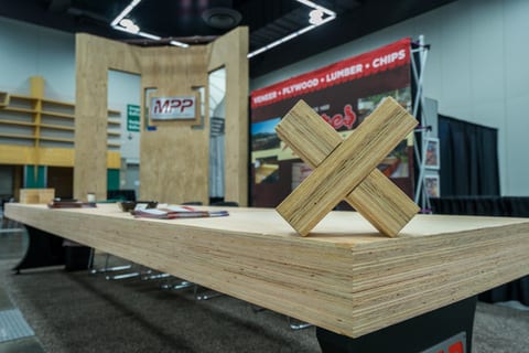 A wooden display table with a large X-shaped wooden structure on top stands prominently at a Mass Timber Conference trade show booth.