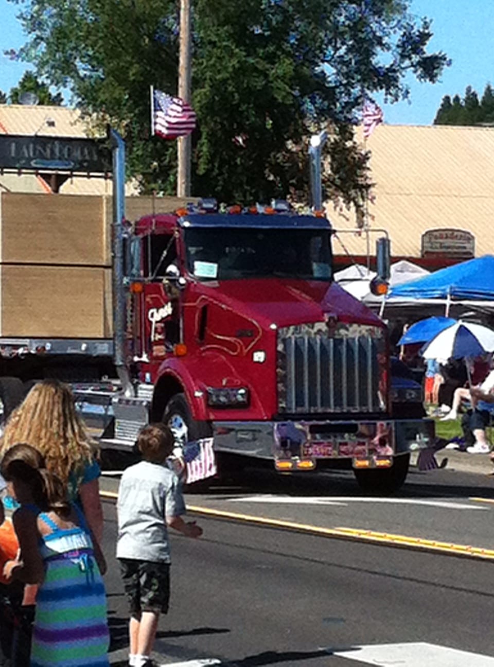 A red semi truck from Freres Engineered Woodmpany, decorated with American flags, drives in a parade, surrounded by people on the street.