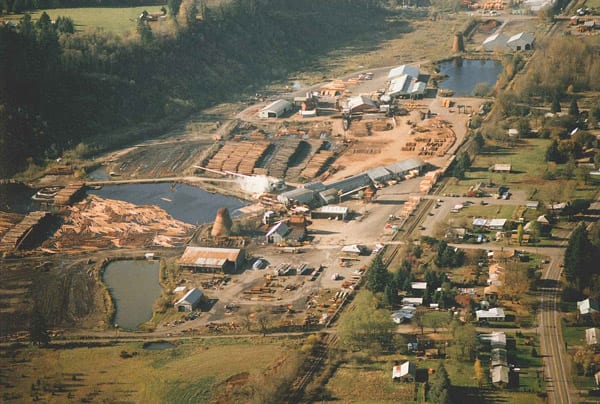 Aerial view of a plywood mill with stacks of logs, log production facilities, ponds, and nearby houses.