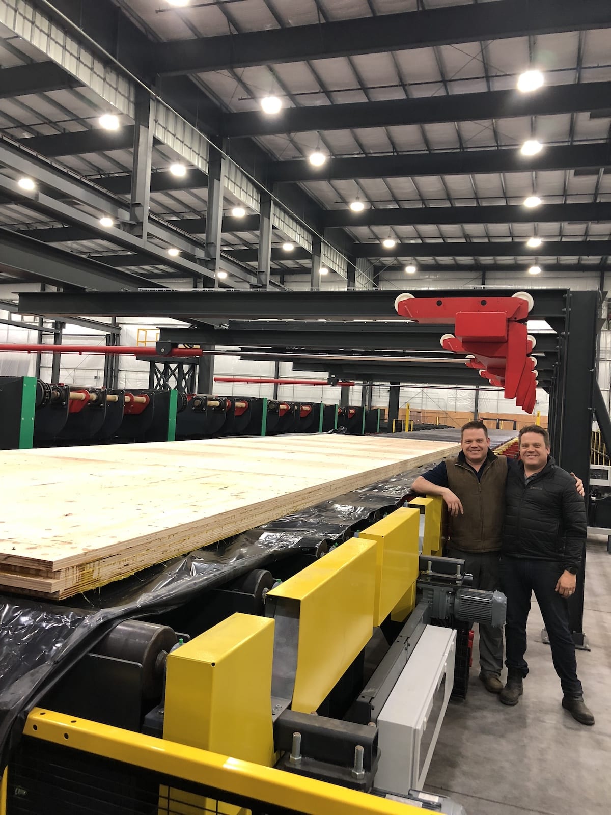 Two people stand by a large Mass Ply Panel processing machine inside an industrial warehouse or factory.