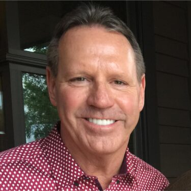 Rob Freres smiling portrait in a red patterned shirt stands outdoors near a building with glass windows.