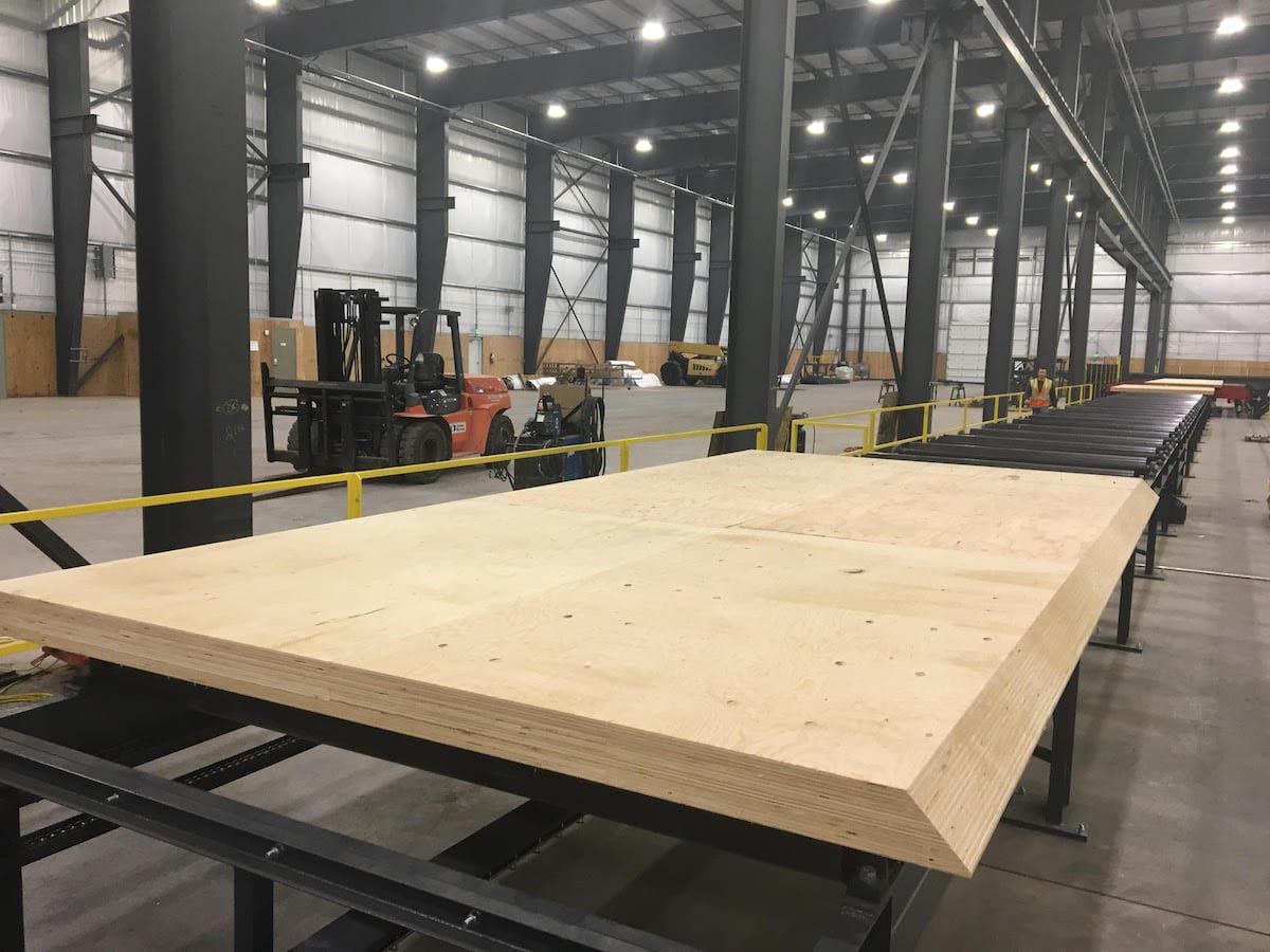 Large sheet of engineered wood on a conveyor in a spacious warehouse, with forklifts and industrial equipment in the background.