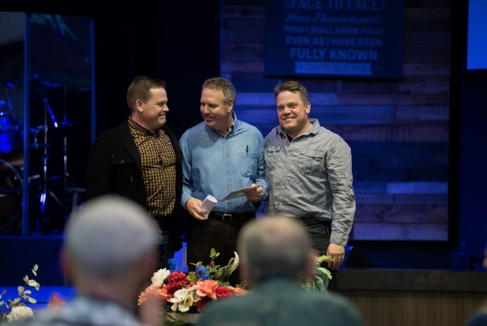 Three men stand together smiling on a stage in Stayton, with people seated and flowers in the foreground.