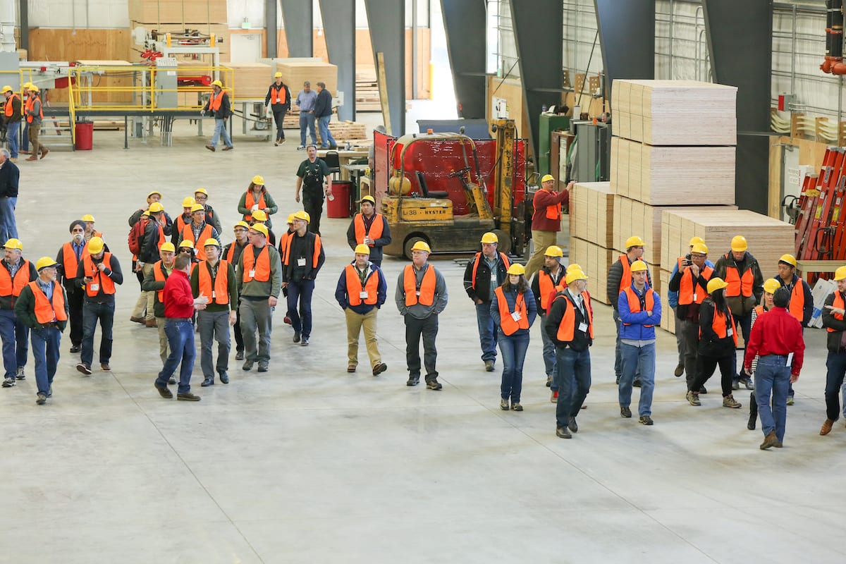 A group of people in orange safety vests and yellow hard hats tour a large industrial warehouse with stacked wood during the Mass Timber Conference.