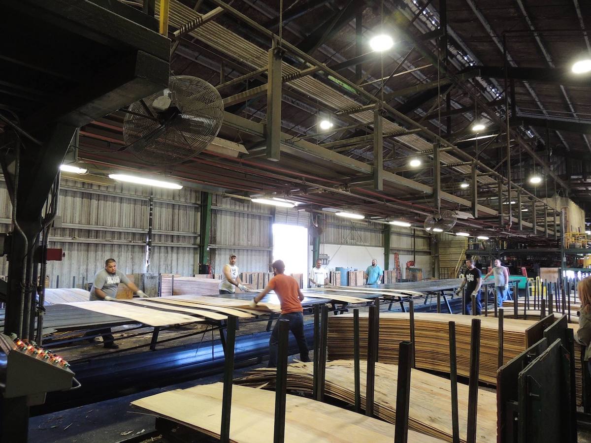 Workers in a large factory move and sort sheets of engineered wood inside an industrial warehouse.