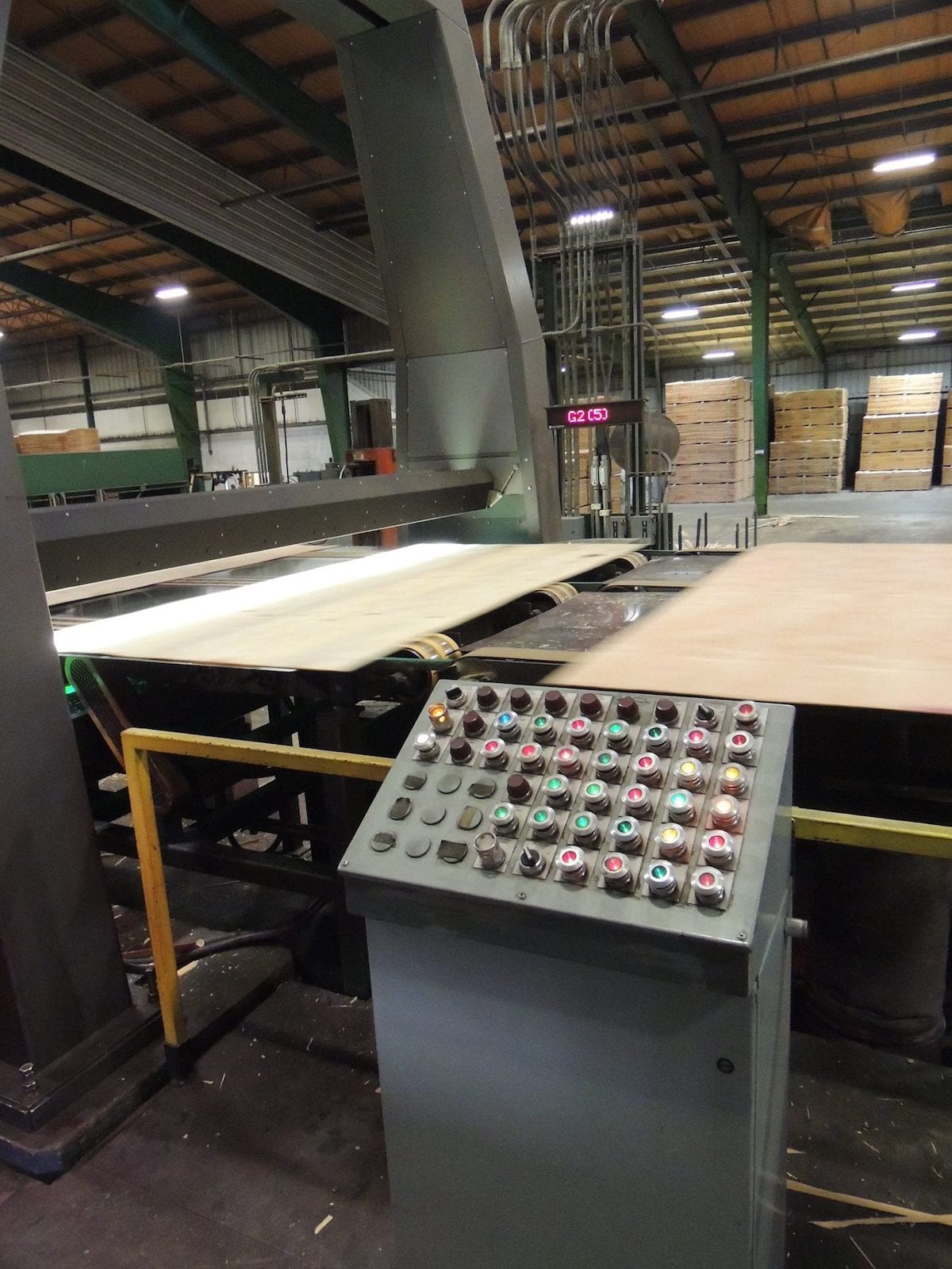 Control panel and machinery in a factory with stacked wood sheets, industrial equipment, and insights into the dynamic in the background.