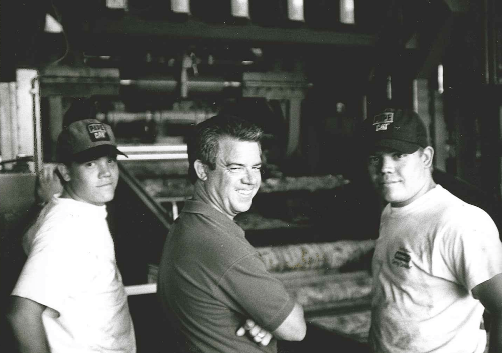 Three men in casual clothing and hats, including Ted Freres, stand smiling inside an industrial or factory setting.
