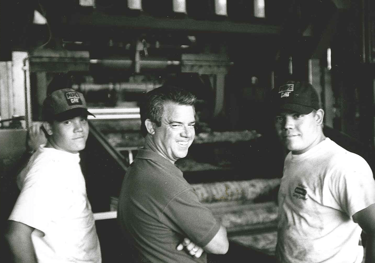 Three men in casual clothing and hats, including Ted Freres, stand smiling inside an industrial or factory setting.