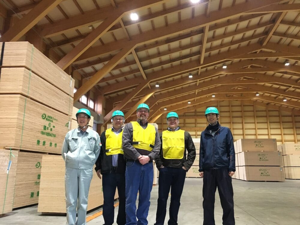 Five people in hard hats stand inside a warehouse, surrounded by stacked plywood and wooden beams—an active scene in the Wood Products Industry.
