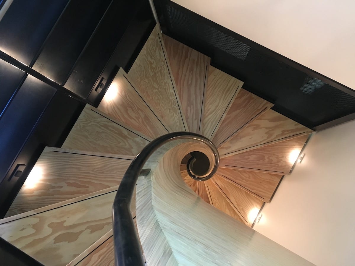 Spiral staircase crafted from engineered wood, featuring lights on each step, viewed from above.