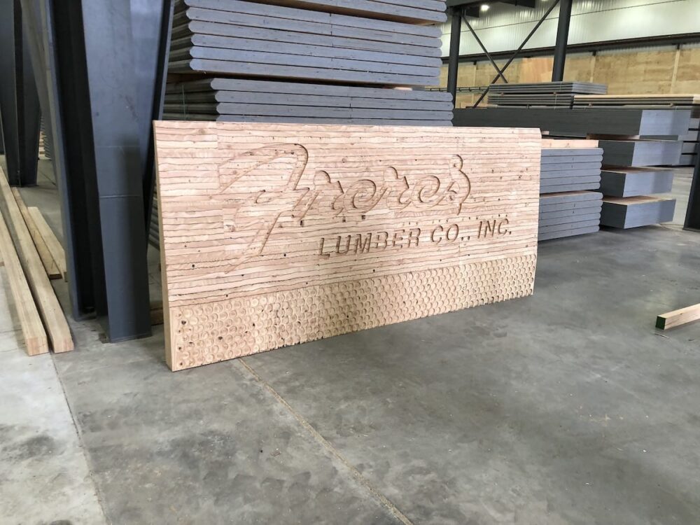 Large wooden sign with Freres Engineered Wood., Inc. and APA Certified engraved, leaning against stacked wood in a warehouse.