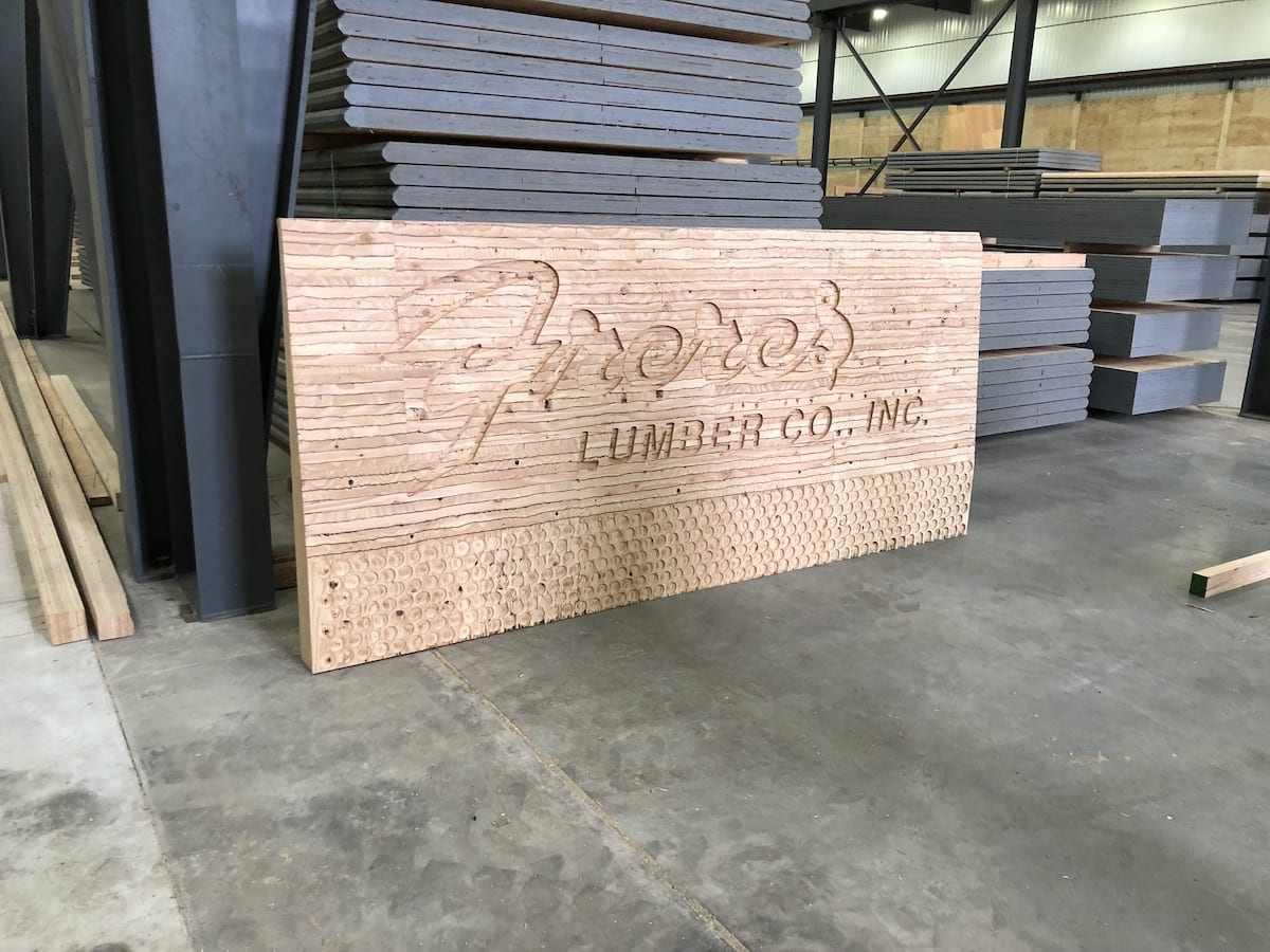 Large wooden sign with Freres Engineered Wood., Inc. and APA Certified engraved, leaning against stacked wood in a warehouse.