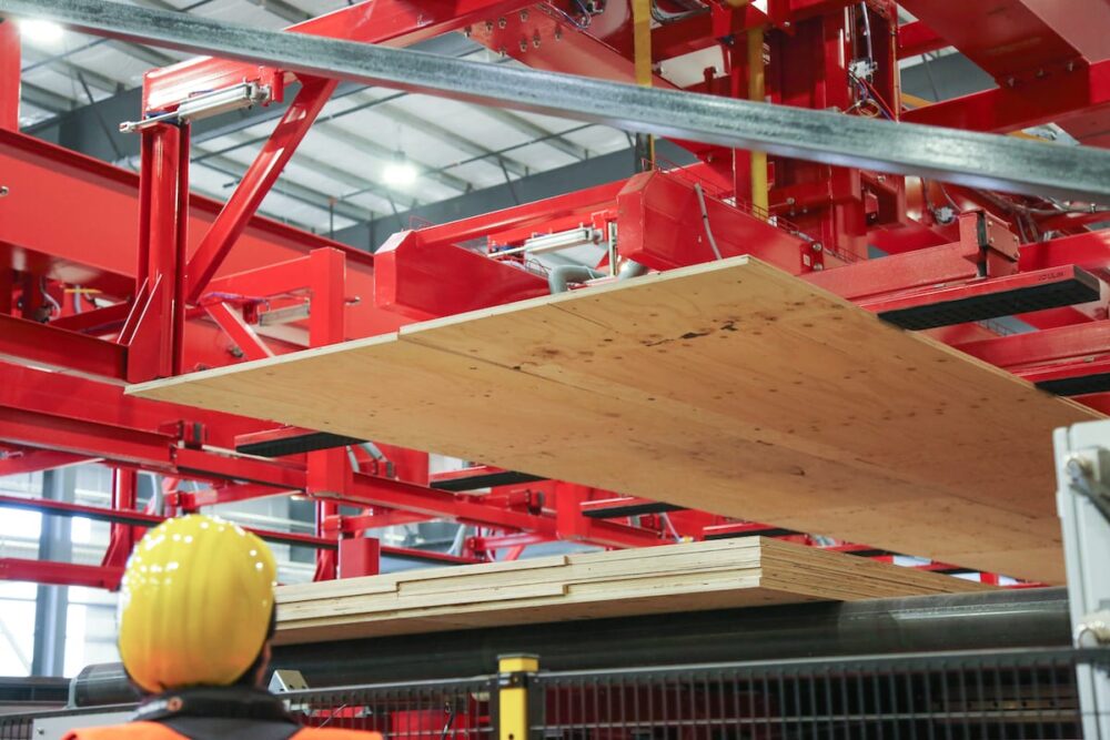A worker watches a machine lift a large wood panel in a factory with red metal structures.