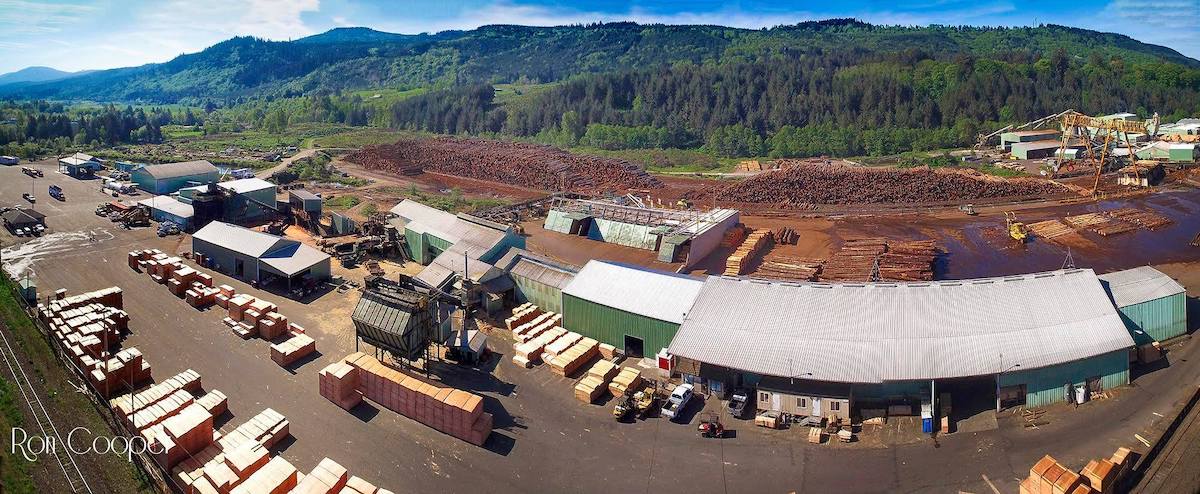 Aerial view of Western Plywood’s lumber yard with stacks of wood, warehouses, and forested hills in the background.