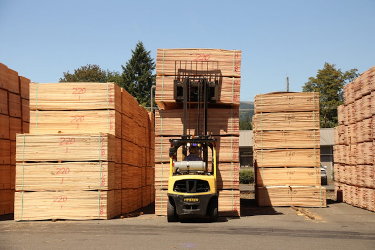 A yellow forklift lifts a stack of lumber between large piles of wooden planks in a lumberyard.