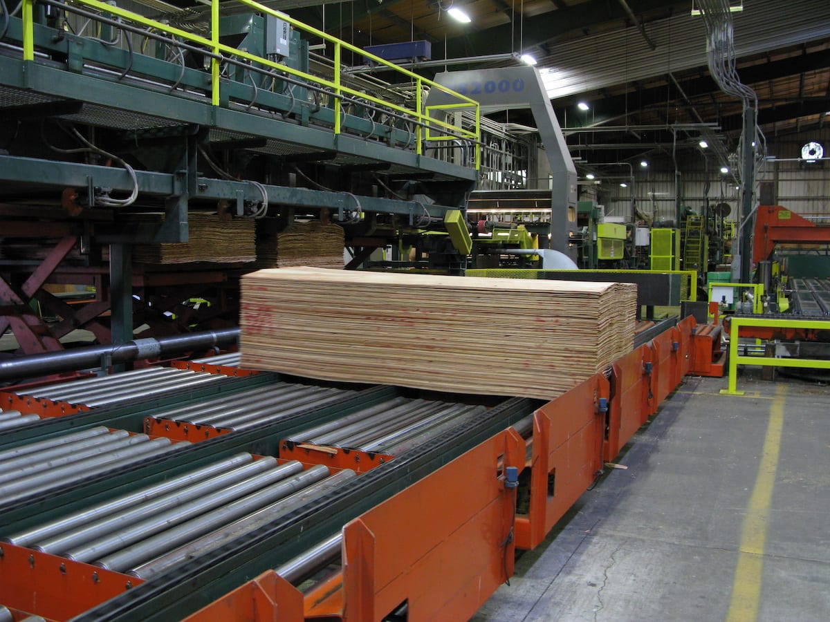 A stack of plywood sheets on a conveyor belt in a large industrial manufacturing facility.