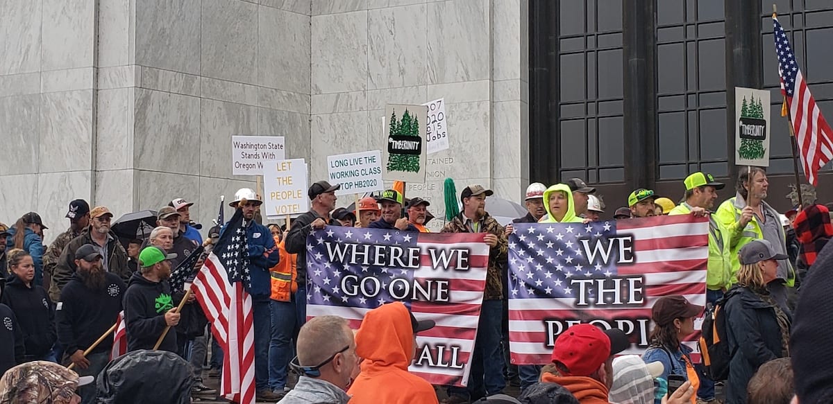 Crowd holding American flags and protest signs during a public rally outside a government building.
