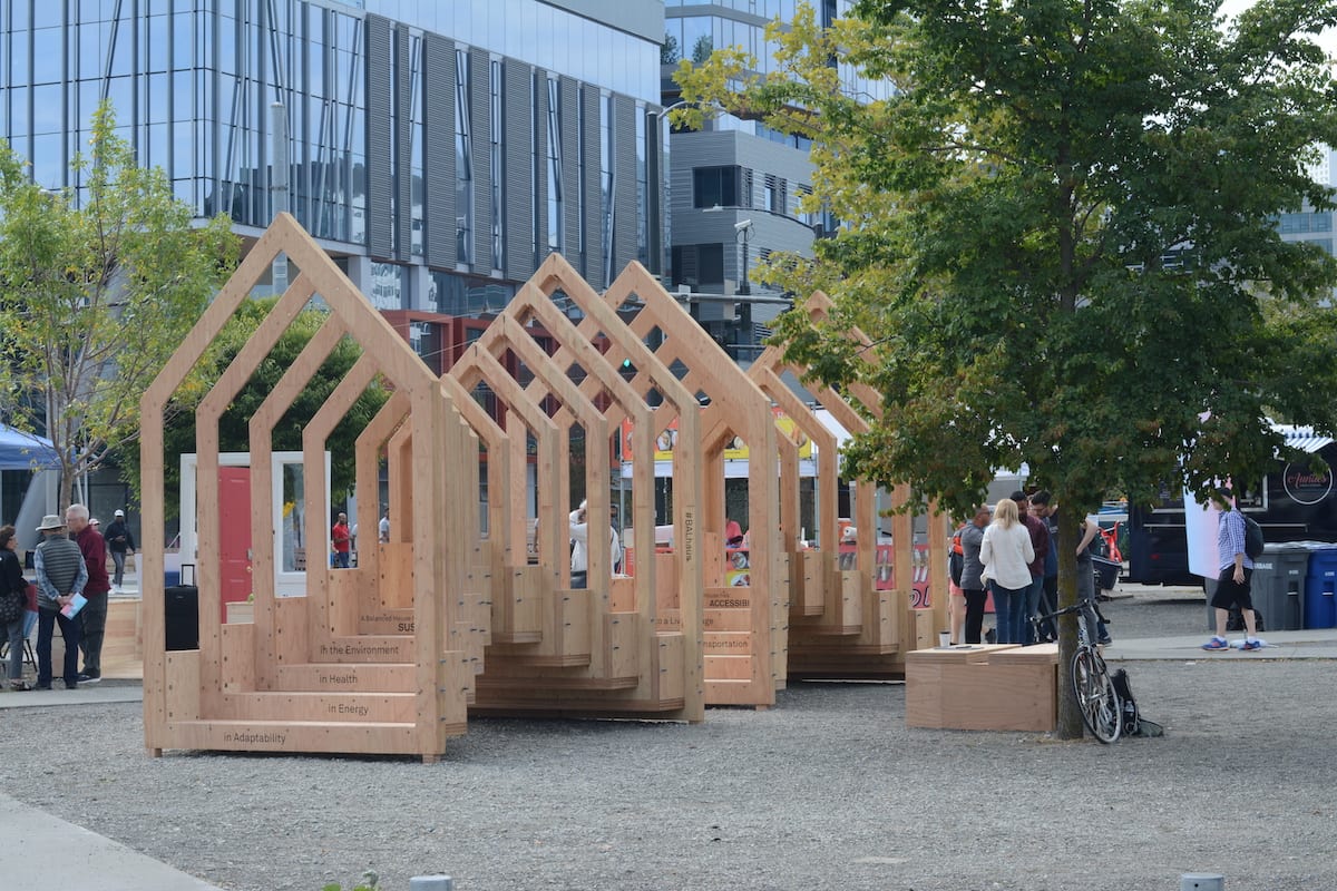 Open-frame wooden house structures displayed in an urban plaza with people and trees nearby.