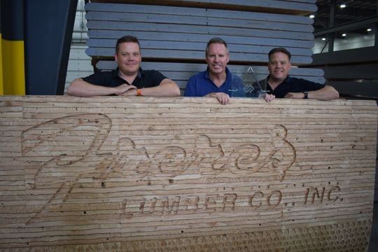 Three men stand behind a large stack of lumber engraved with Frederick Lumber Co., Inc. inside a warehouse.