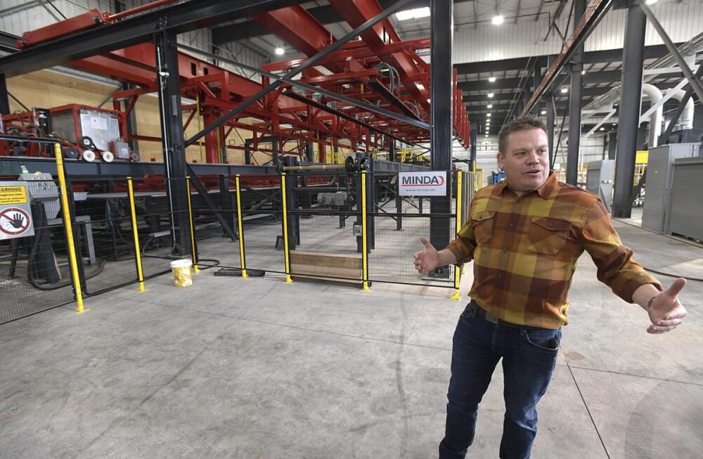 A man in a plaid shirt gestures while standing in a large industrial warehouse with red machinery.