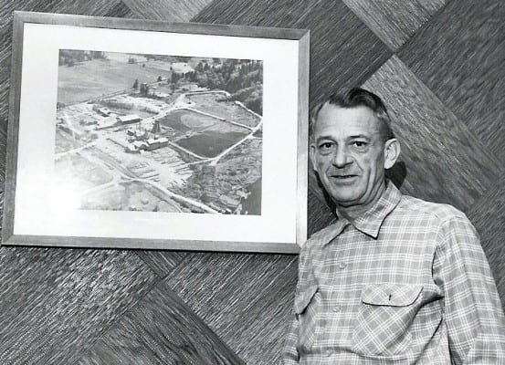 Man in a plaid shirt standing next to a framed aerial photo by T.G. Freres on a wood-paneled wall.