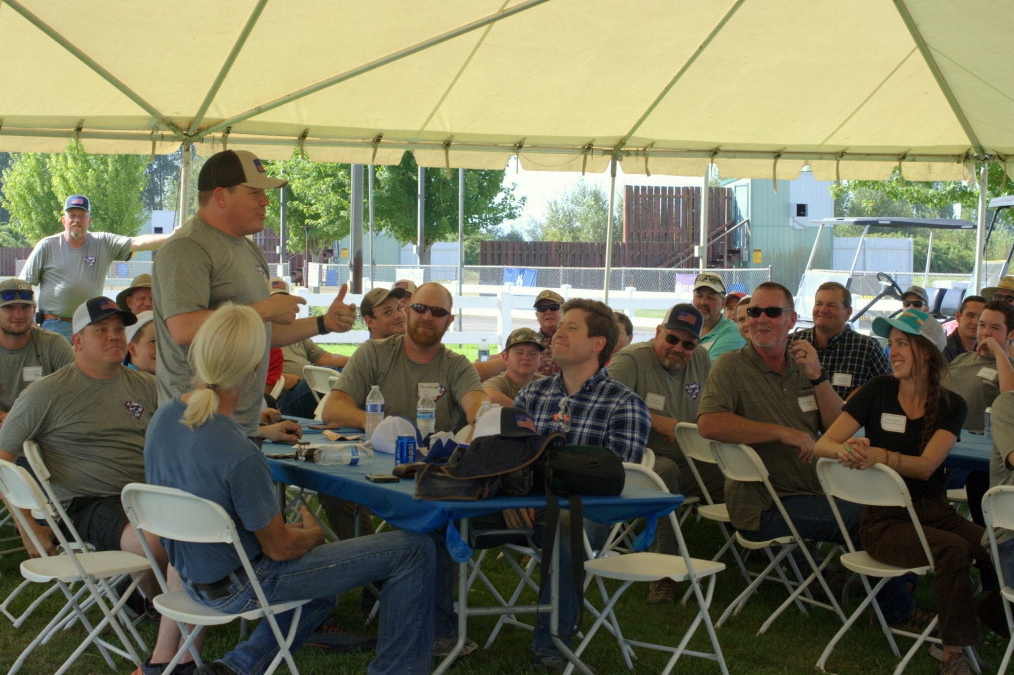 A man speaks to a seated group under a large tent at the Classic Sporting Clay Invitational outdoor gathering.