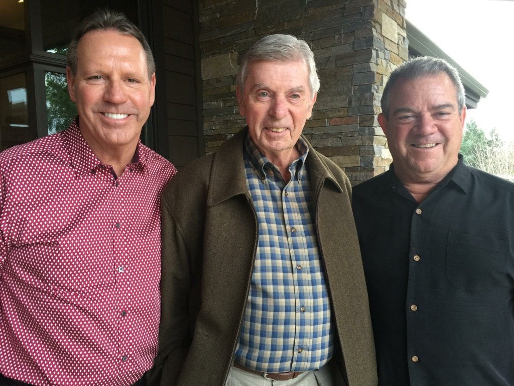Three smiling men, including Bob Freres, stand together in front of a brick building, posing for a photo.