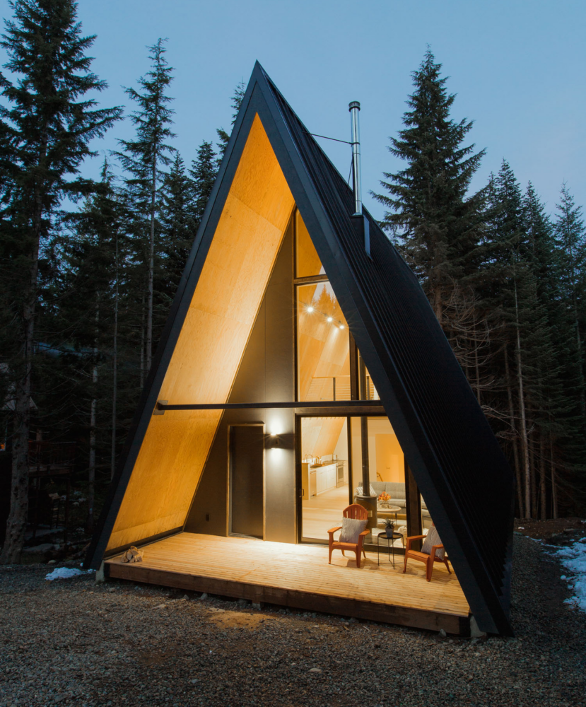 A modern A-frame cabin with large glass windows, glowing warmly, surrounded by tall pine trees at dusk.