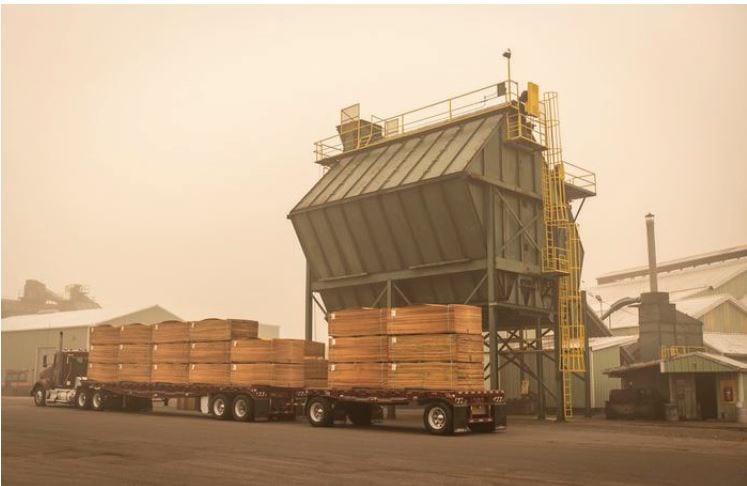 A flatbed truck loaded with stacks of lumber is parked by an industrial building on a foggy day.