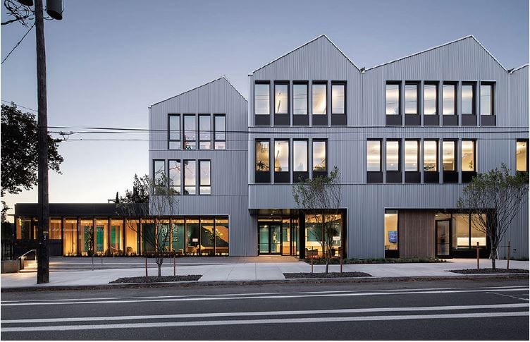 Modern three-story building with large windows and sharp rooflines at dusk, facing a street.