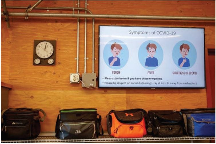 Wall clock, screen showing COVID-19 symptoms, and several lunch bags lined up on a shelf below.