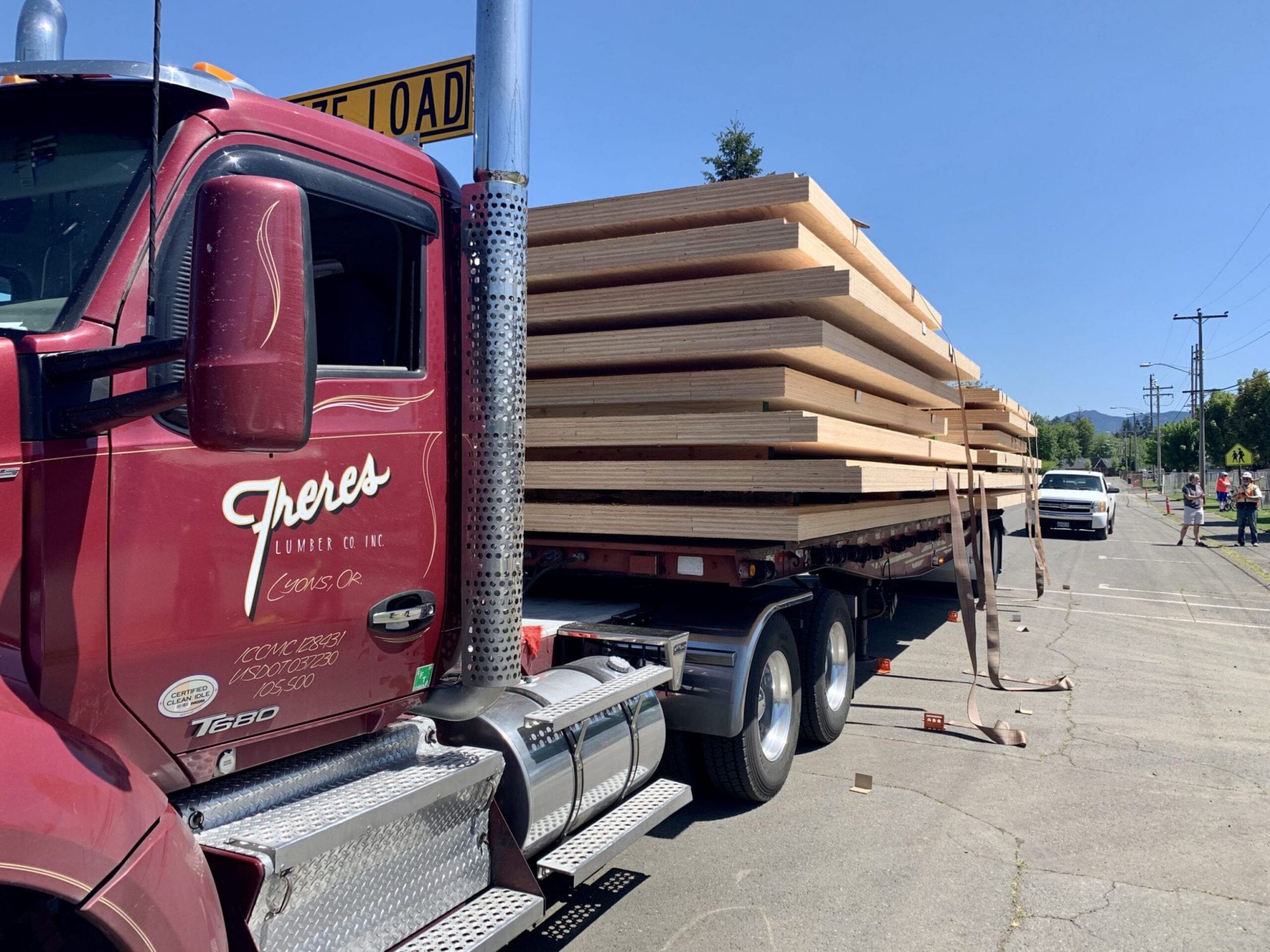 A red semi-truck loaded with large lumber planks is parked on a sunny street near a crosswalk, delivering materials for new school construction.