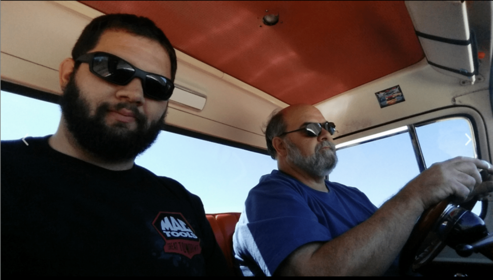 Two men with beards and sunglasses sit in a car; one is driving while the other, Bob Trusty, relaxes in the passenger seat.