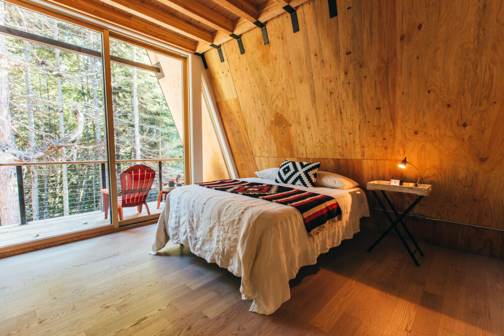 A cozy wood-paneled bedroom with a bed, a lamp, and large window overlooking trees and a red chair on the balcony.