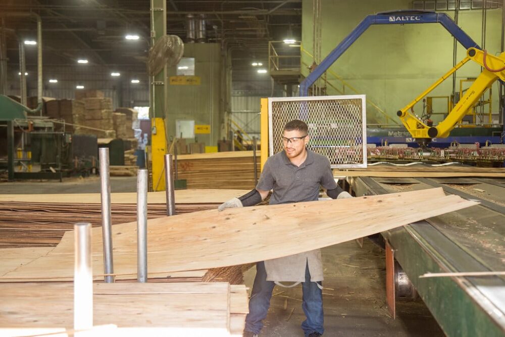 A man wearing safety glasses handles a large wooden sheet in a factory with industrial equipment.