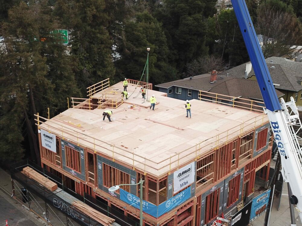 Construction workers install wooden flooring using mass ply panels on the upper level of a building under construction, near tall trees.