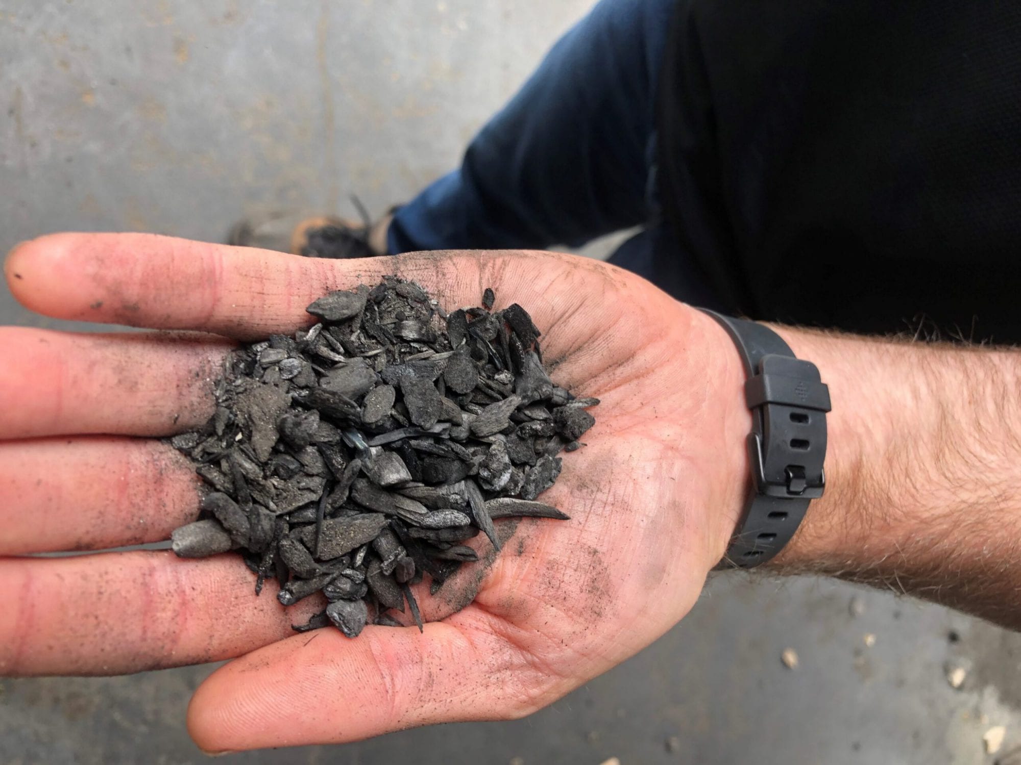 A hand holding a pile of small, black biochar pieces over a gray surface.