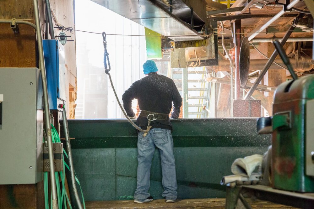 Worker in safety harness stands at industrial site, complying with vaccine executive order, facing large window with bright light streaming in.