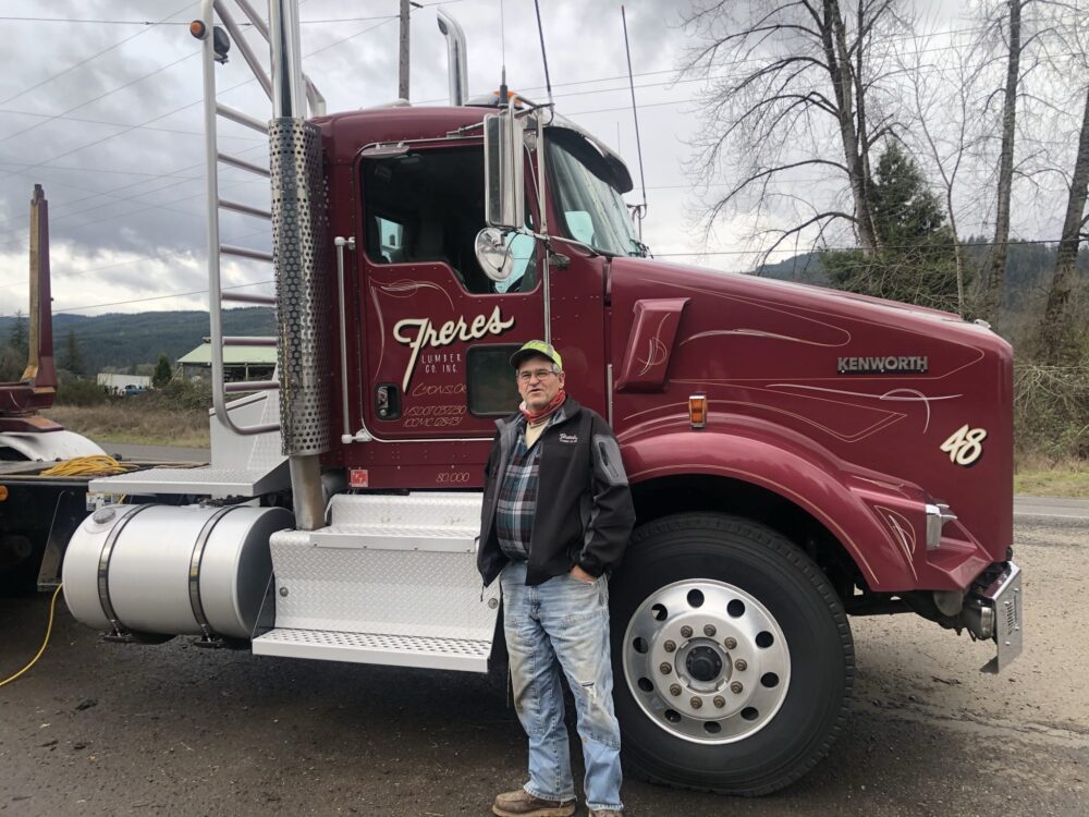 A man in a jacket and cap stands by a maroon Kenworth truck on a cloudy day near trees.