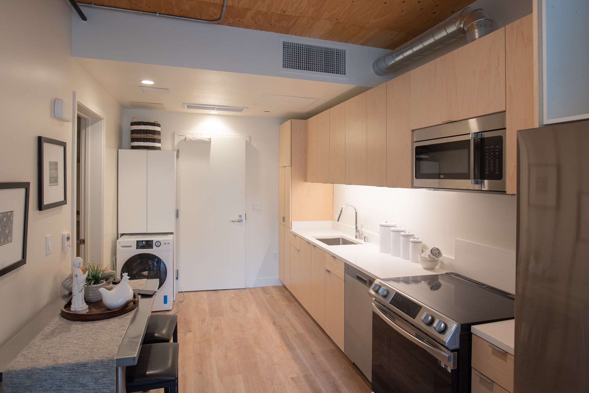 Modern kitchen with light wood cabinets, stacked washer and dryer, and stainless steel appliances.