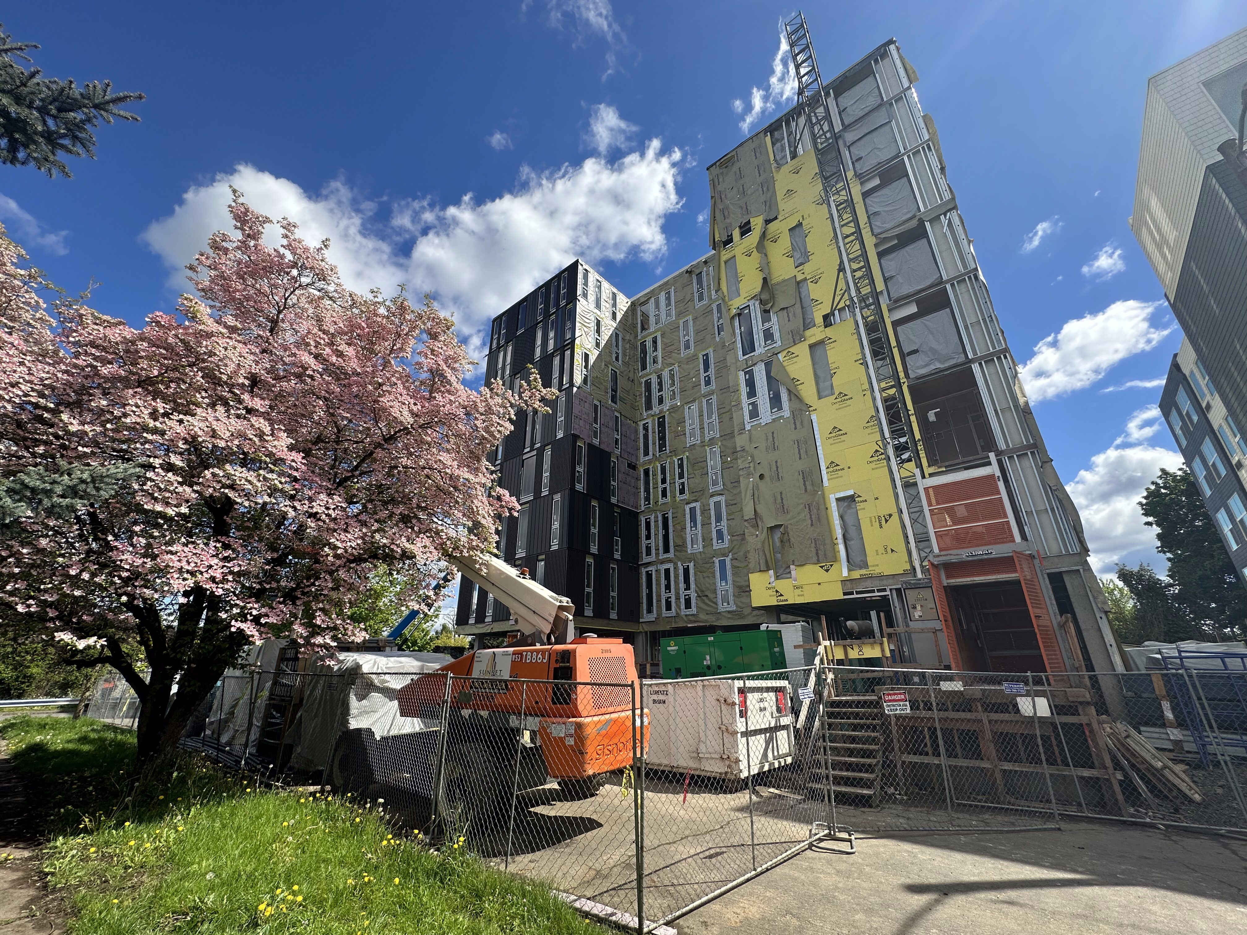 A modern building under construction beside a blooming tree under a blue sky with clouds.